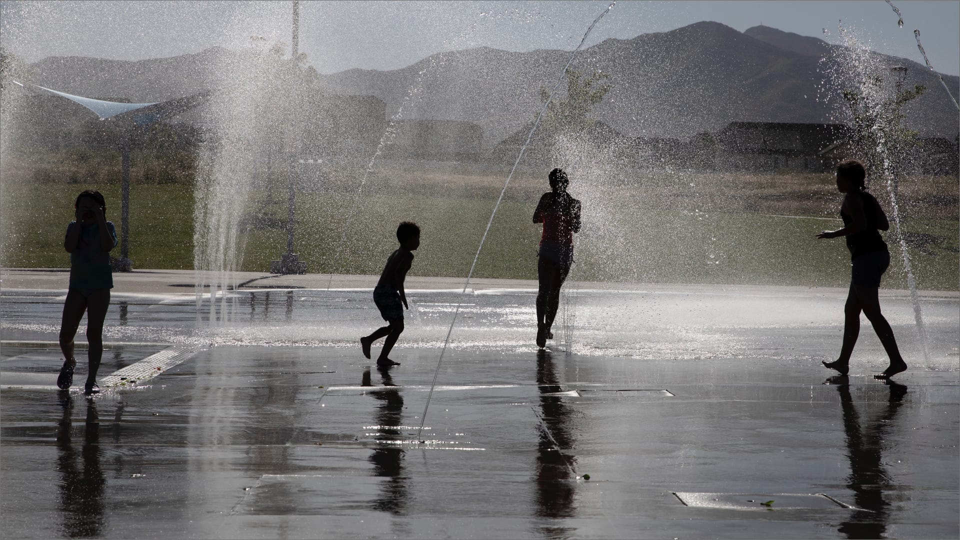 Children playing in a splash pad with water jets shooting up from the ground. The scene is backlit, casting shadows and creating silhouettes. There are mountains and grass fields in the background under a clear sky.