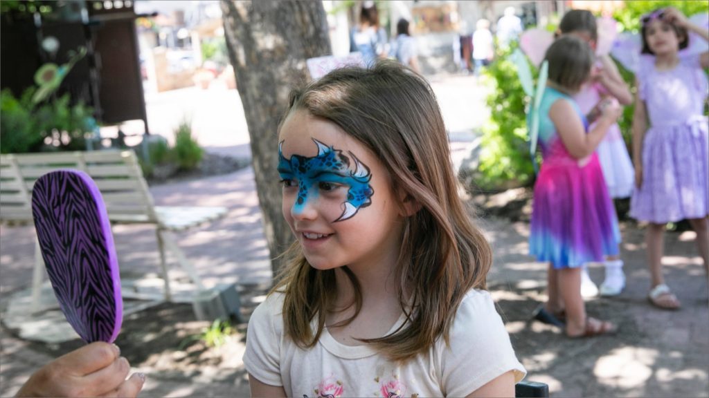 A young girl with colorful face paint smiles while looking in a handheld mirror. She stands outdoors in a park, surrounded by other children in colorful clothing. Trees and benches are visible in the background.
