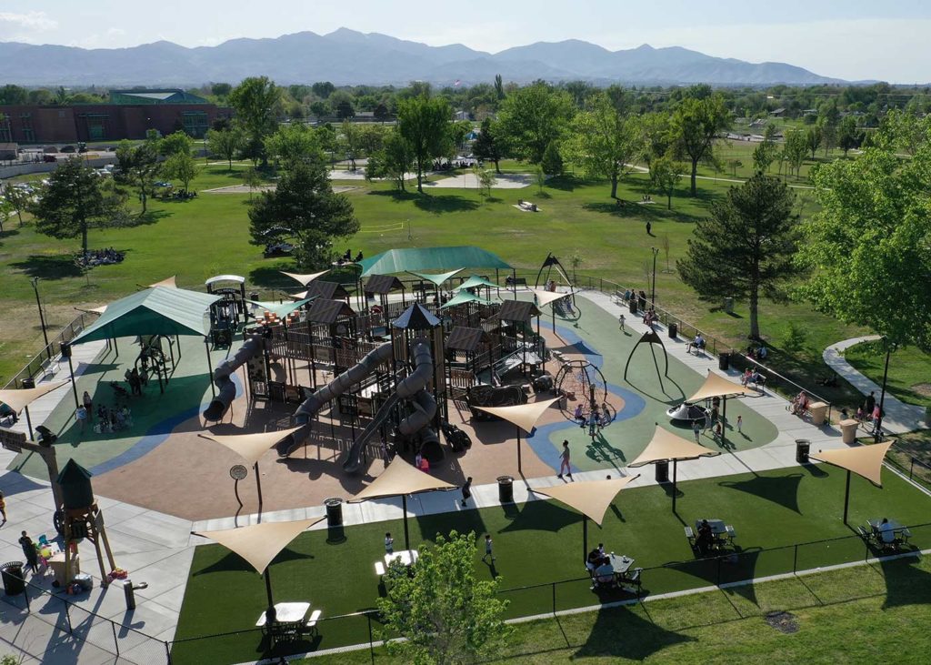 Aerial view of a large playground in a park, featuring slides, climbing structures, and swings. Surrounding the playground are trees, grass, and walking paths. Mountains are visible in the background under a clear blue sky.