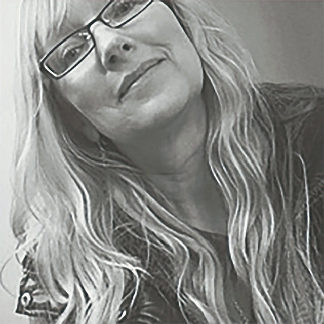 A grayscale photo of a person with long, wavy hair wearing glasses and a denim jacket. They are tilting their head slightly and smiling softly at the camera.