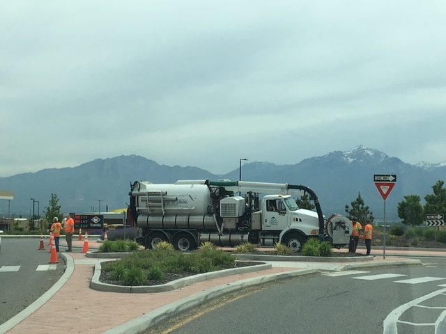 A utility truck with a large vacuum tank is parked at a roundabout. Several workers wearing orange vests and hard hats are nearby, with traffic cones around them. Mountain peaks can be seen in the background under a cloudy sky.