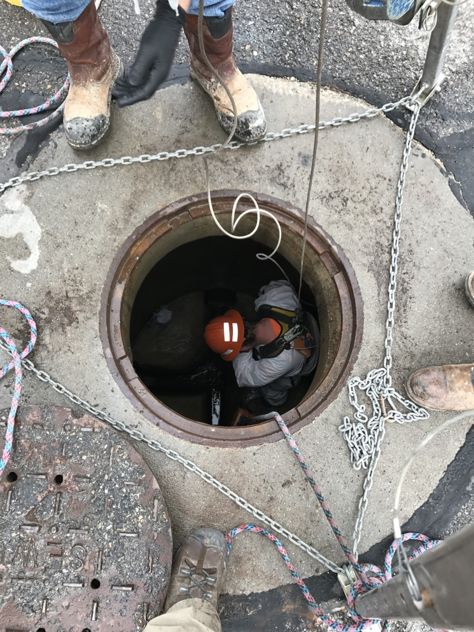 A worker in protective gear and helmet is climbing out of a manhole. Ropes and chains are arranged around the opening. Several people stand nearby, visible from the knees down, wearing work boots.