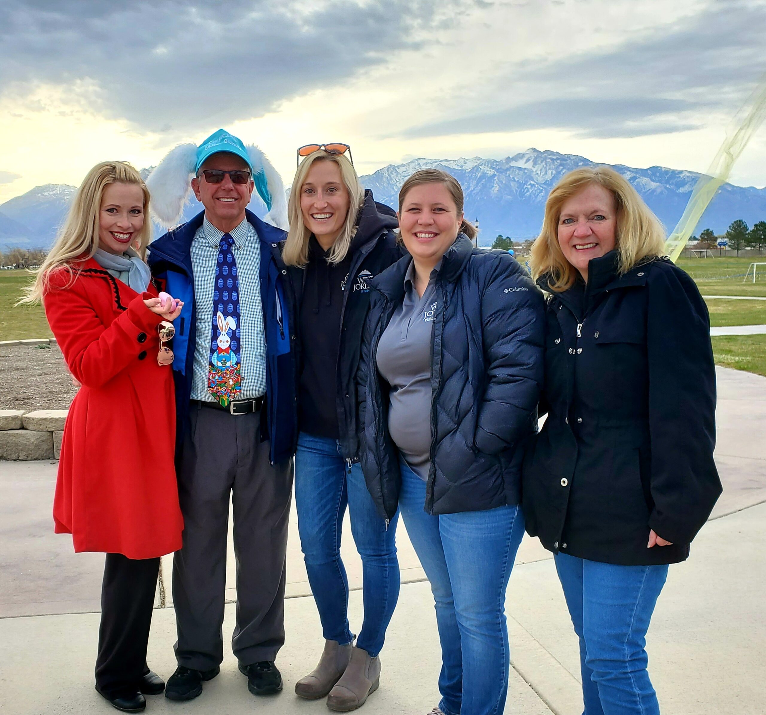 A group of five people standing outdoors, smiling at the camera. One person is wearing a Santa hat and tie. Mountains and a cloudy sky are in the background. Everyone is dressed in winter clothing.