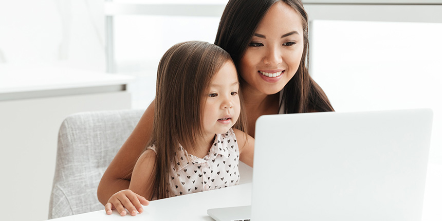 A woman and a young girl sit at a table, smiling and looking at a laptop screen. The woman has long dark hair, and the girl has shoulder-length hair. They appear engaged and happy in a bright room with natural light.