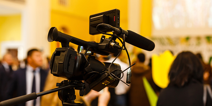 A video camera on a tripod is set up in a crowded event space, focused on a presentation. People are gathered in the background, with yellow walls and decorations adding a bright atmosphere.
