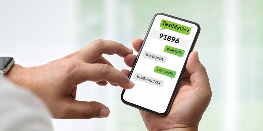 A person holding a smartphone displaying the TextMyGov app. The screen shows a number 91896 and buttons labeled WICOUNCIL, WIJEVENTS, and WIJNEWSLETTER for text updates. The background is softly blurred.
