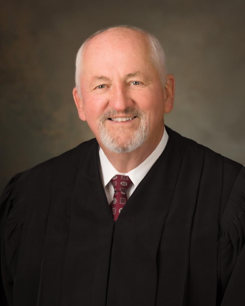 A smiling man with white hair and a beard is wearing a black judges robe and a red patterned tie. The background is a neutral color with a soft, blurred texture.