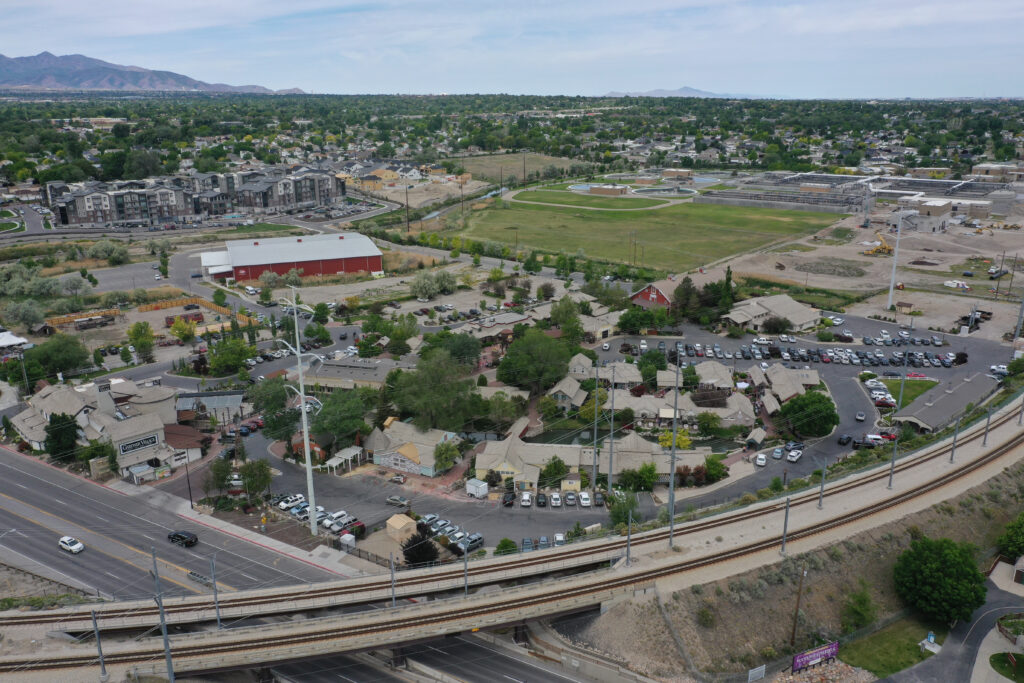 Aerial view of a suburban area featuring a curving road, train tracks, residential buildings, and parking lots. In the background, theres an open green space and distant hills under a partly cloudy sky.