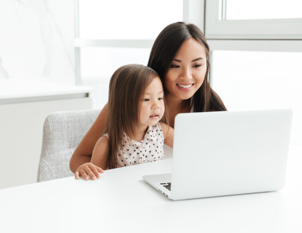 A woman and a young girl sit together at a white table, both smiling and looking at a laptop. They appear to be in a bright room with large windows.
