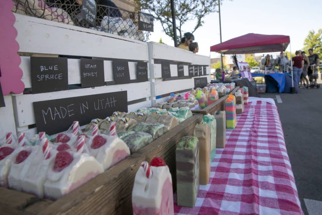 A display of colorful artisanal soaps on a table with a red and white checkered cloth at an outdoor market. Signs indicate various scents like blue spruce and Utah honey. Other market stalls and people are visible in the background.
