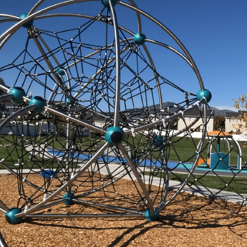 A modern playground features a large geometric climbing structure made of metal bars and ropes. The structure sits on woodchip ground with grass and pathways visible in the background under a clear blue sky.