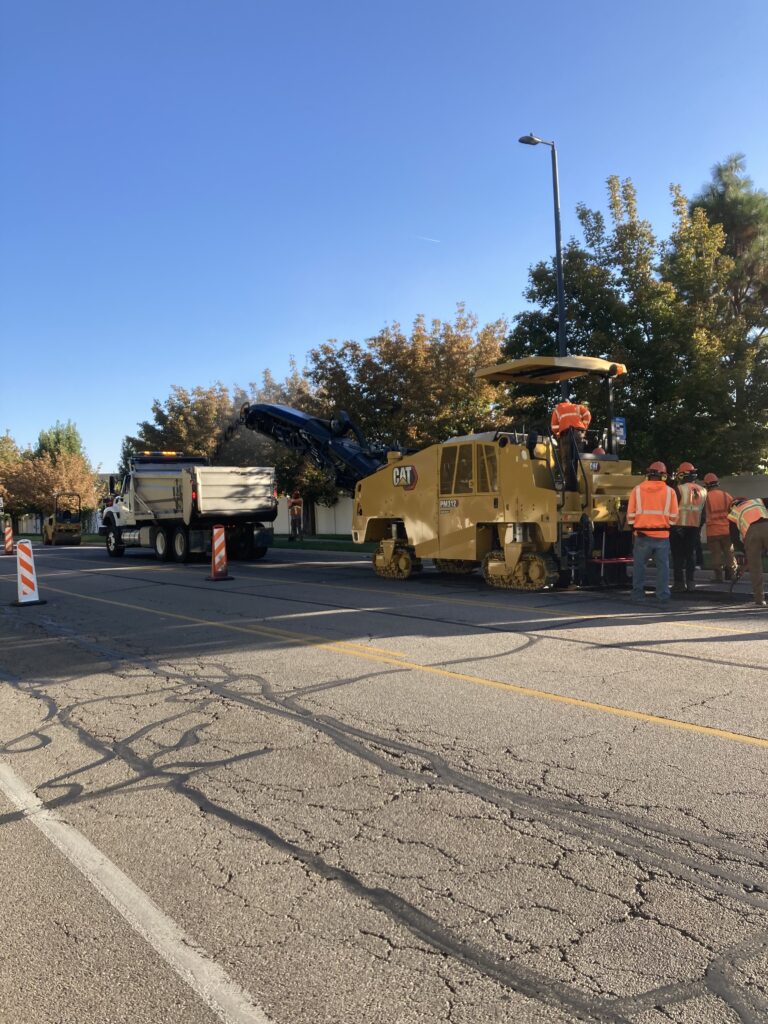 Roadwork scene with construction workers in orange vests operating heavy machinery on a sunny day. A large yellow asphalt paver and a dump truck are on the road. Traffic cones and cracked pavement are visible, with trees in the background.