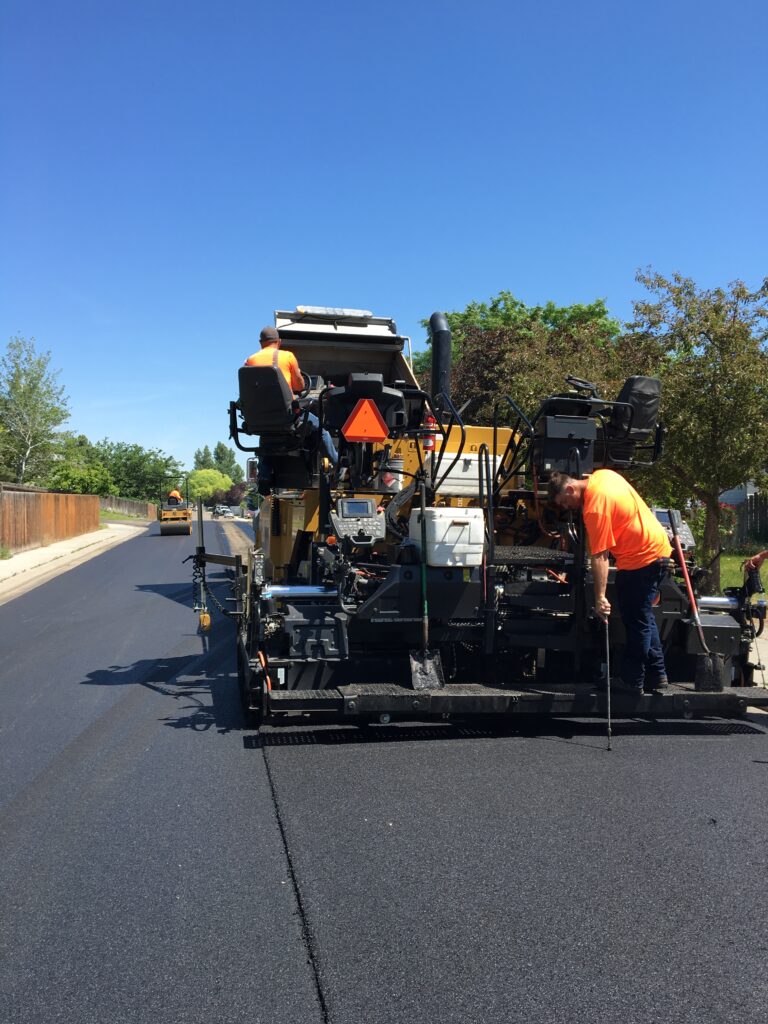 Workers in bright orange shirts operate a paving machine on a freshly laid asphalt road under a clear blue sky. Trees and a wooden fence line the street, and a smaller construction vehicle can be seen in the background.