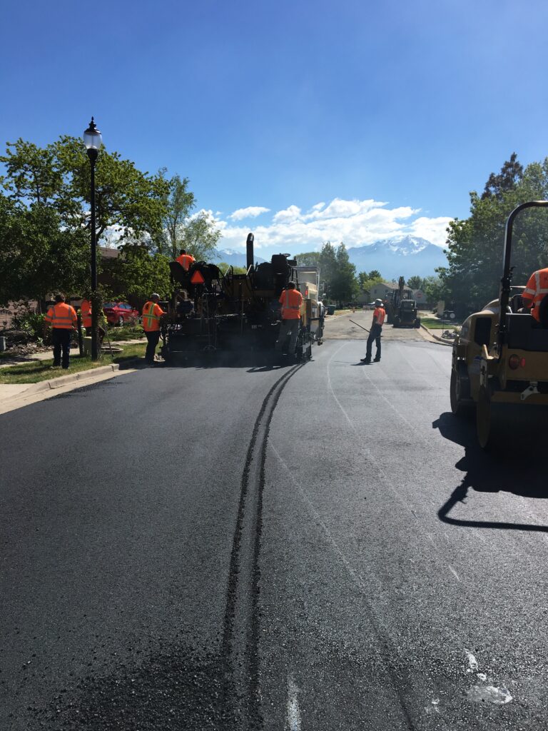 Workers in orange vests paving a sunny road with construction equipment. Trees and a lamppost line the street, with mountains visible under a clear blue sky.