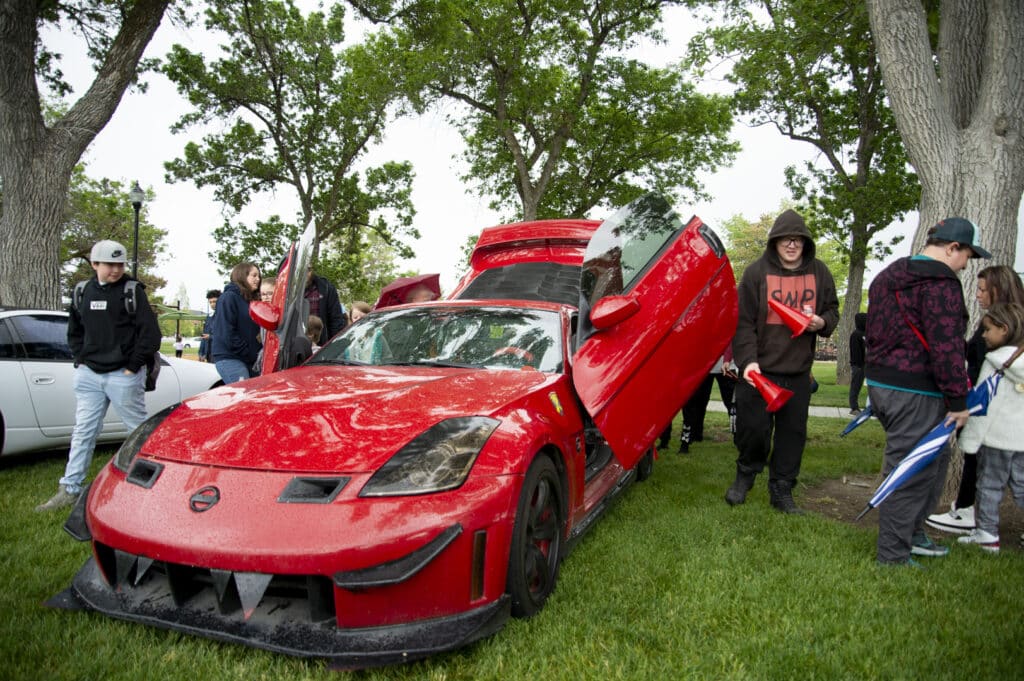 A shiny red sports car with vertical doors open is displayed on grass. Several people, including a person with a black hoodie, are gathered around, admiring it. The scene is set under leafy trees with overcast weather.