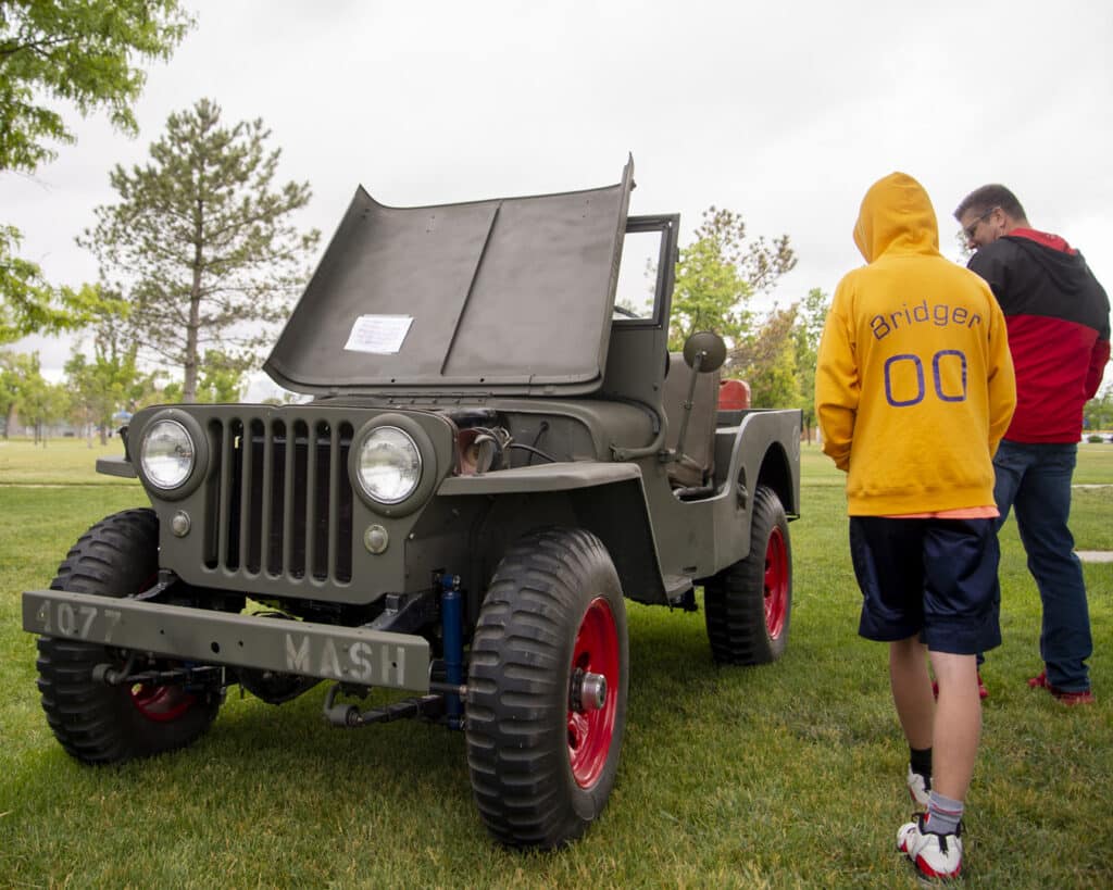 A vintage military jeep with its hood open is displayed on a grassy area. A boy in a yellow Bridger 00 hoodie and a man in a red and black jacket stand nearby, observing the vehicle. Trees and cloudy sky are in the background.