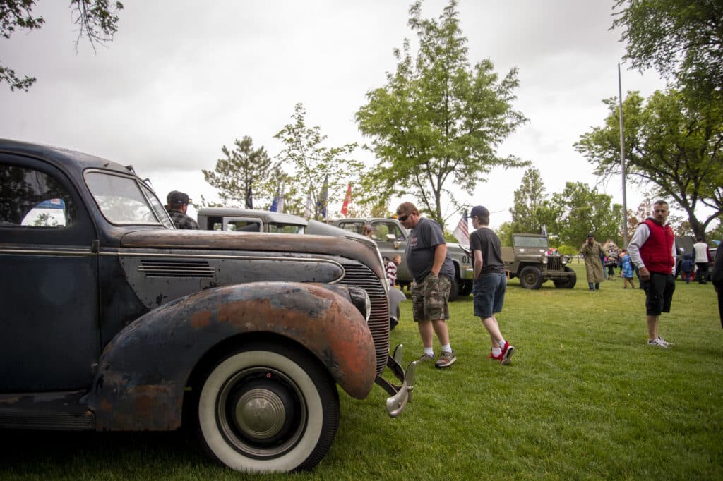 A group of people walk past vintage cars at an outdoor car show on a grassy field. Trees and clouds fill the background. Some attendees are casually dressed and observe the vehicles.
