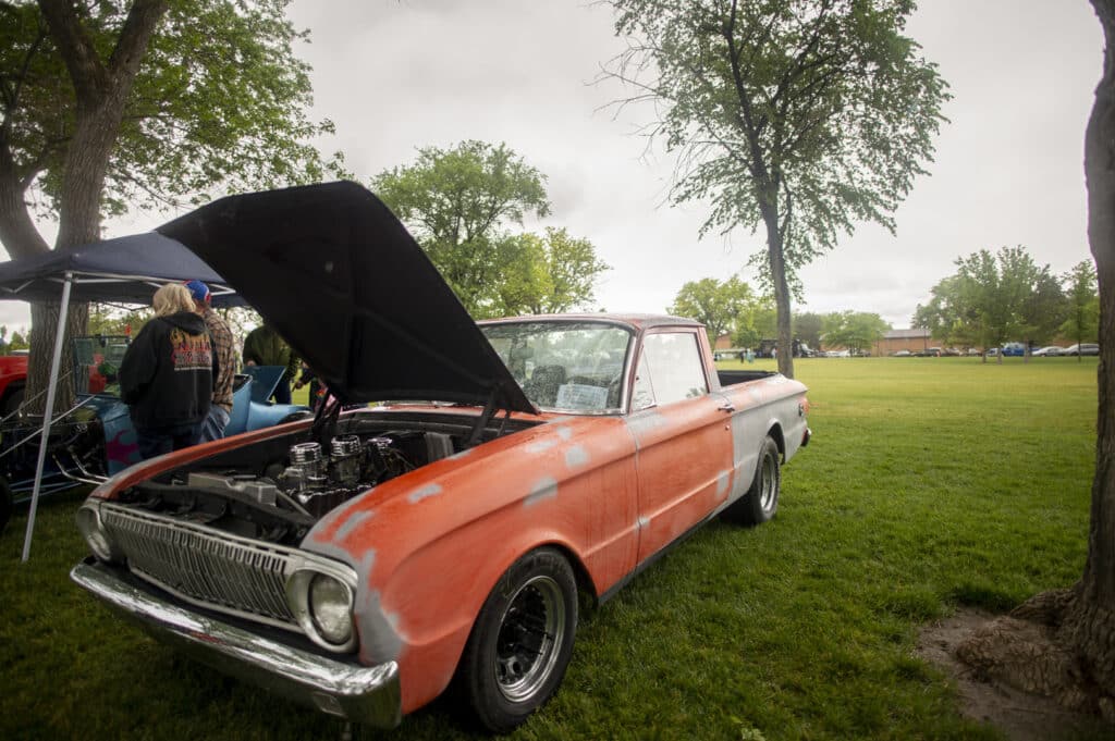 An old orange car with its hood open is parked on a grassy field under cloudy skies. People are gathered under a tent nearby, and trees surround the area.