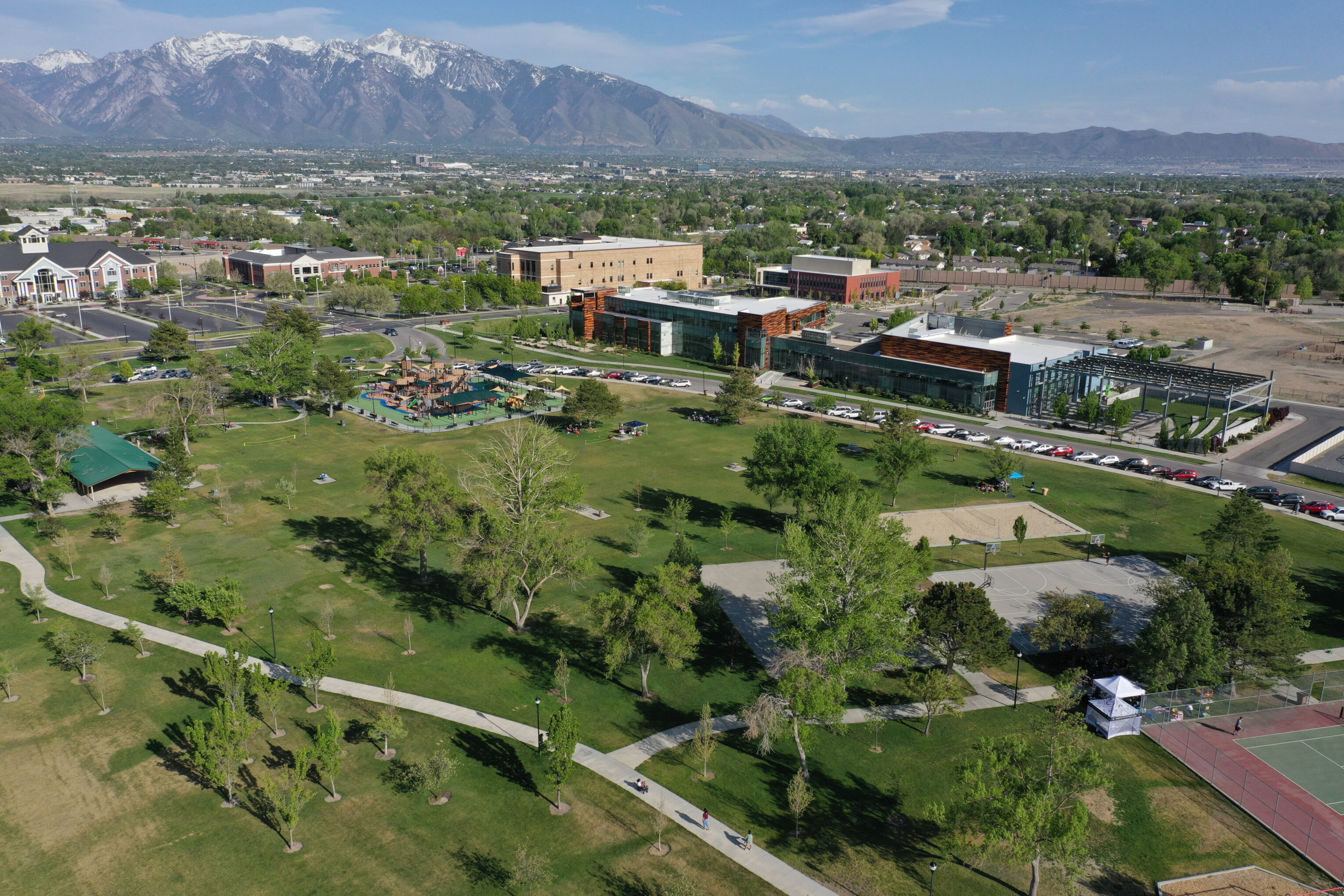 Aerial view of a large park with green lawns, pathways, and trees. There are buildings and parking areas nearby. Snow-capped mountains are visible in the background under a clear blue sky.