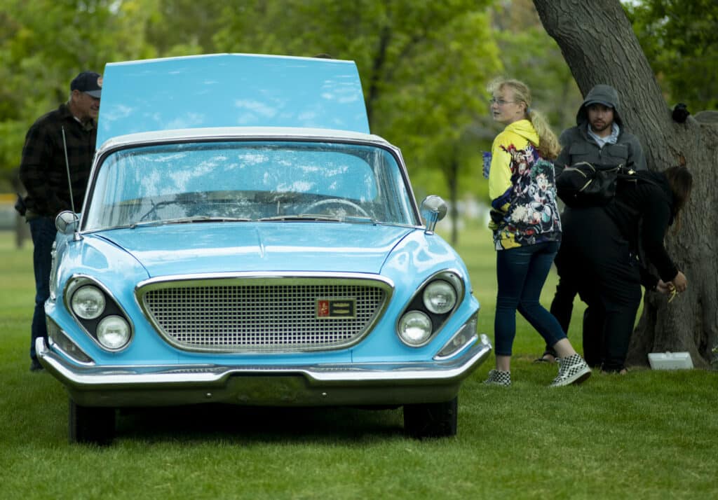 A vintage blue car with its hood open is parked on a grassy area. Four people are nearby, including a woman in a floral jacket and a man by a tree. The scene is set outdoors with trees and a cloudy sky in the background.