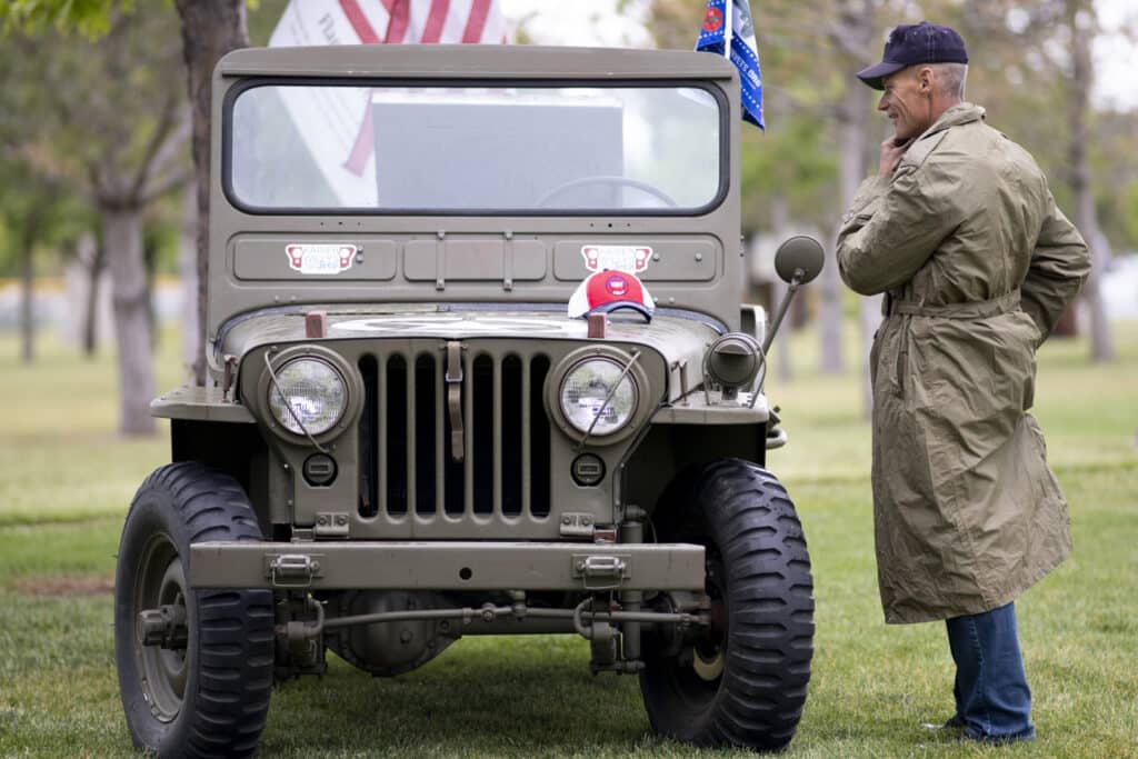 A man in a trench coat stands beside a vintage military jeep in a park. The jeep has an American flag and a cap on the hood. Trees and grass surround the vehicle, and the man appears to be observing the details.