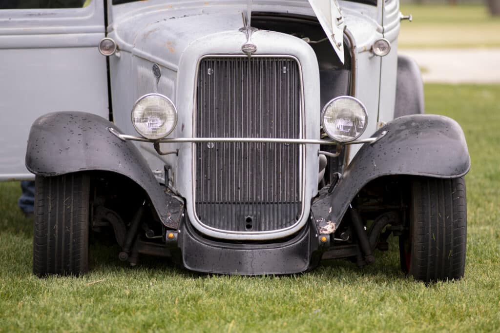 Front view of a vintage car with its hood open, revealing a classic grille and two round headlights. The black fenders contrast with the light-colored body. The car is parked on grass.