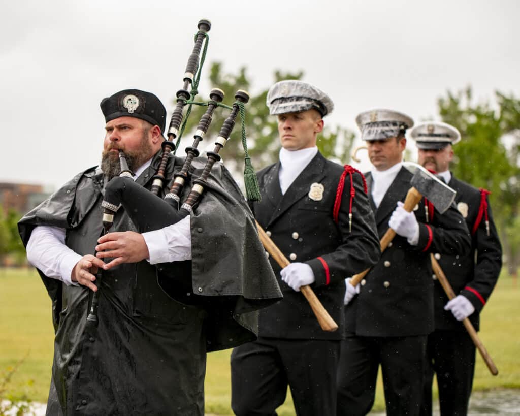 A man in a beret plays bagpipes while three men in formal uniforms, holding ceremonial axes, walk behind him. They are outdoors on a grassy area under a cloudy sky.