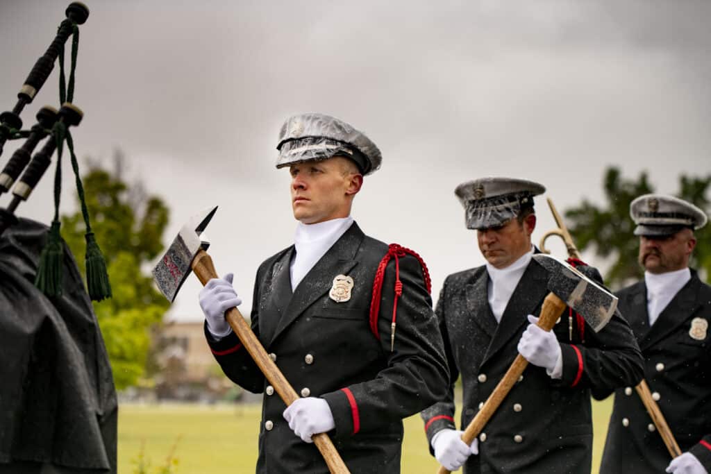Three uniformed individuals march solemnly, each holding an axe. They are dressed in formal black uniforms with white gloves and hats. The sky is overcast, and trees can be seen in the background.