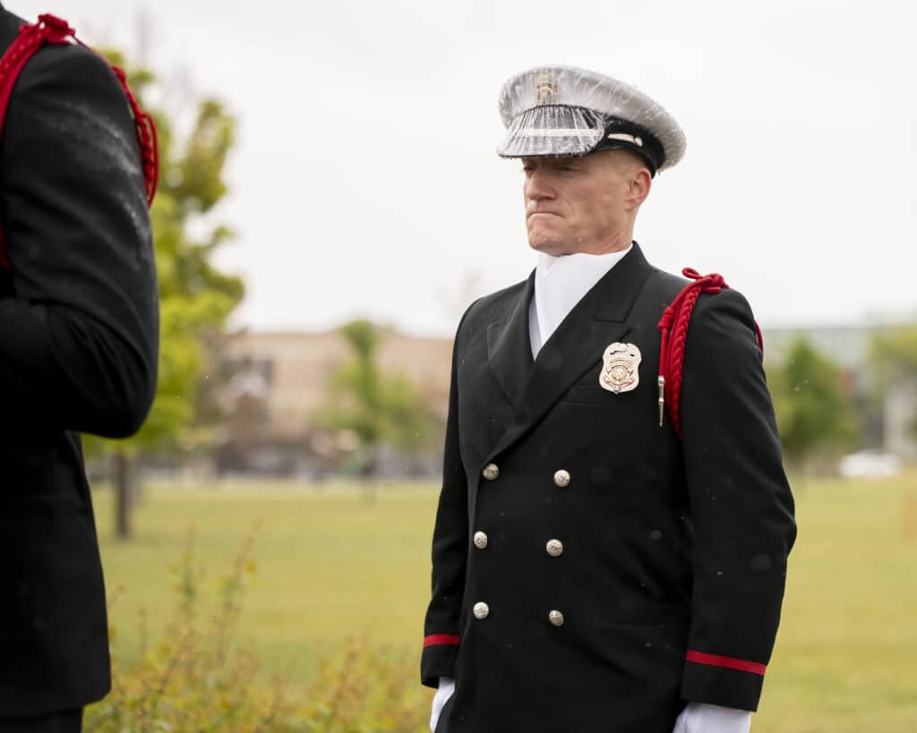 A uniformed officer with a silver cap and badge stands outdoors in front of a grassy field. He is wearing a black coat with silver buttons and red shoulder cords.