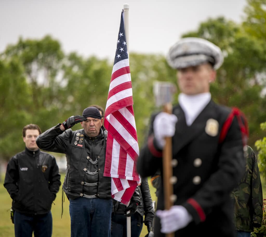 A man in a leather vest salutes the American flag while standing outdoors, surrounded by other people. He appears focused and respectful. A uniformed person stands in the foreground holding a pole. Trees are visible in the background under a cloudy sky.