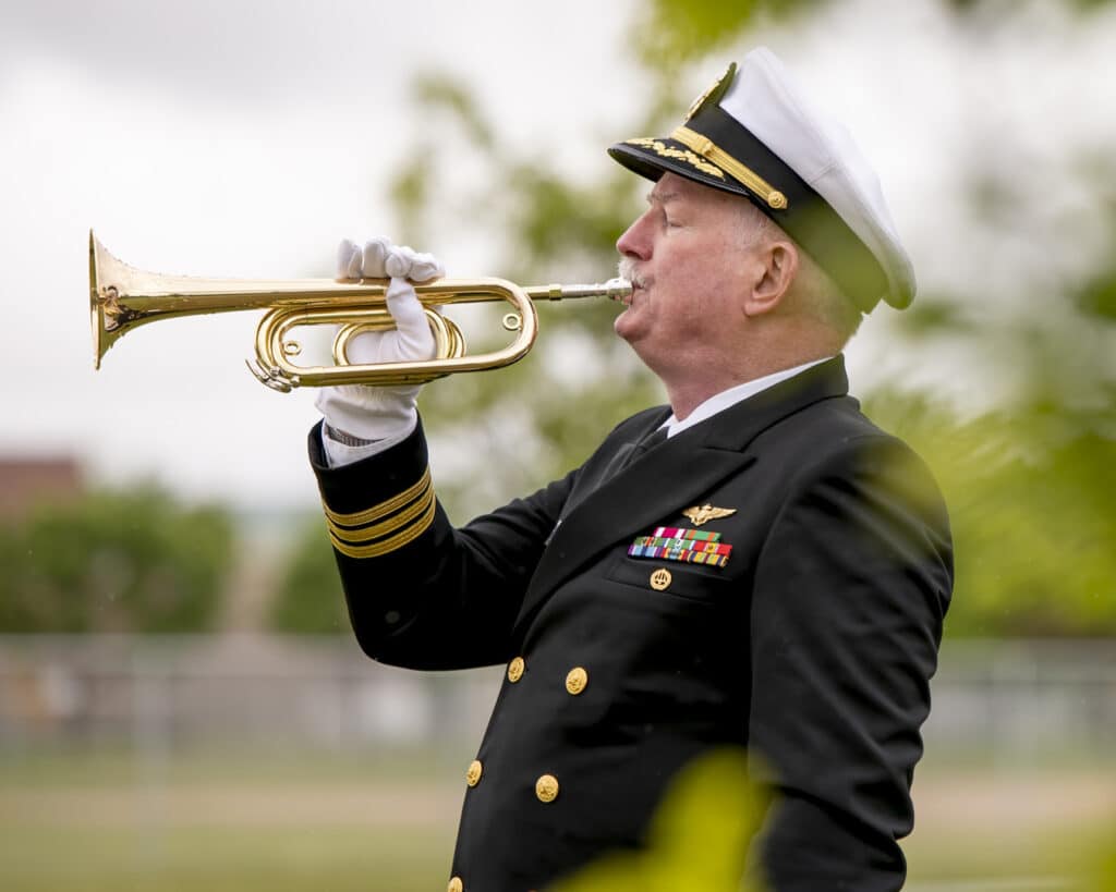 A man in a navy dress uniform plays a brass trumpet outdoors. He wears a white captains hat and white gloves. The background is blurred with green foliage.