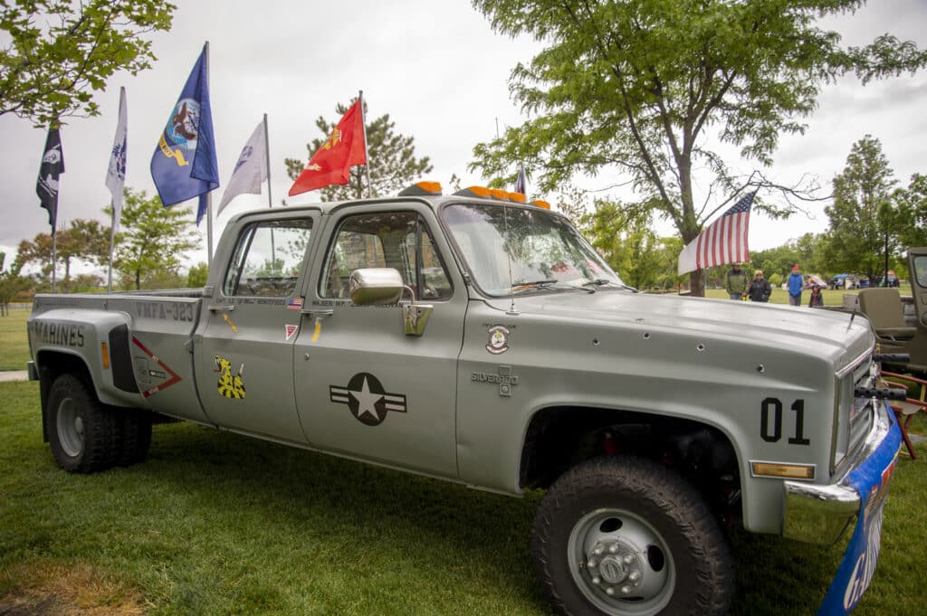 A custom military-themed truck is parked on grass, adorned with various military insignias and decals. Multiple flags, including the American flag, are displayed on poles attached to the truck. Trees and other parked vehicles are in the background.