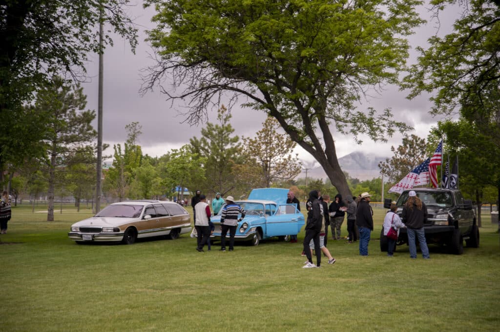 People gather around classic cars at an outdoor car show in a park. The scene includes a blue vintage car with its hood open, flags on a truck, and attendees wearing jackets. Trees and overcast skies are in the background.