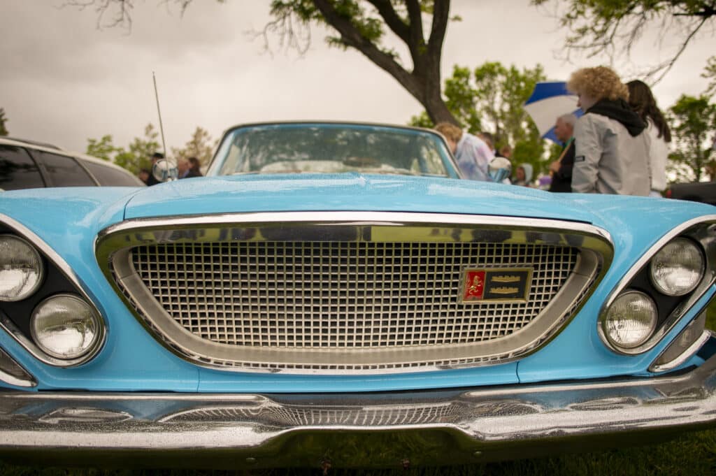 A vintage blue car is shown from the front, displaying a chrome grille with classic styling. A group of people stand in the background, with greenery and a cloudy sky overhead.