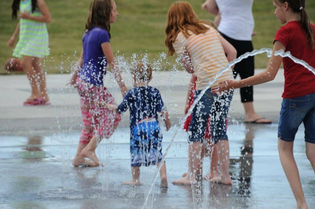 Children play joyfully in a splash pad with water jets on a sunny day. They are barefoot and dressed in colorful summer clothes, running and laughing as water sprays around them. Some adults are nearby, supervising and enjoying the scene.