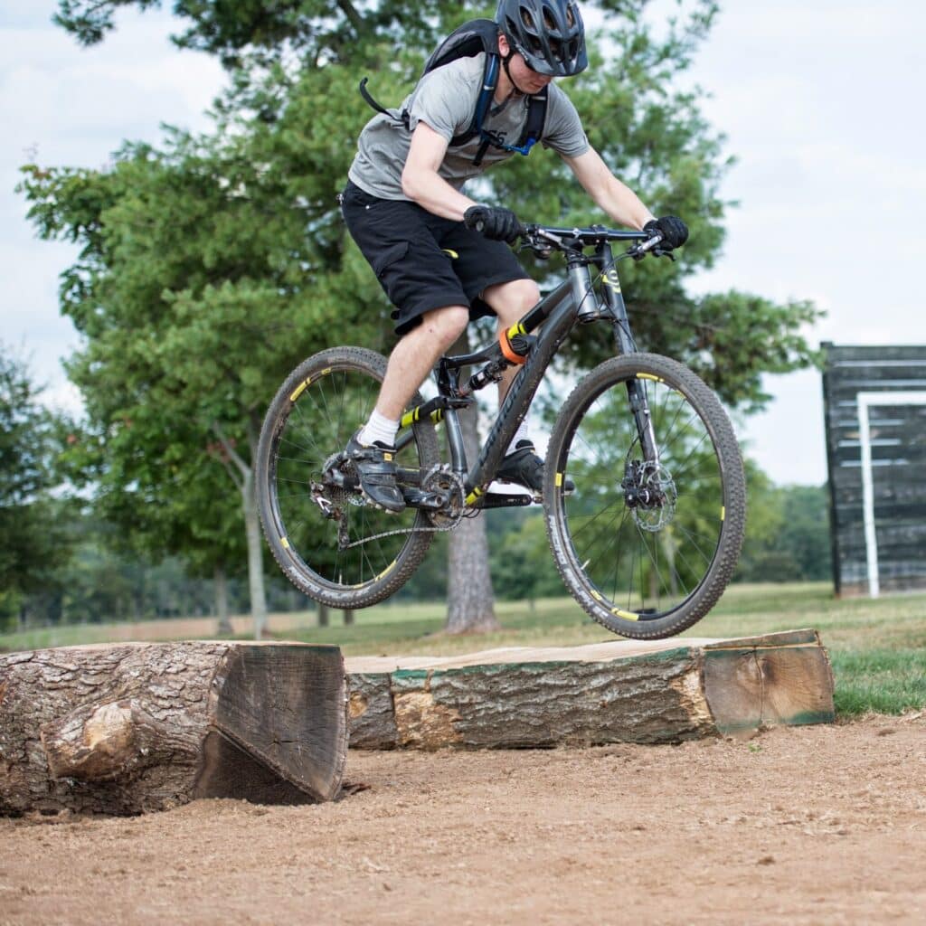 A cyclist wearing a helmet and backpack is performing a jump over a wooden obstacle on a trail. The background features trees and grassy areas under a partly cloudy sky.