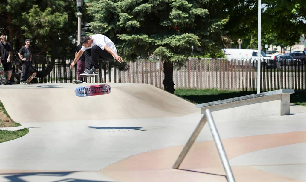 A skateboarder mid-air performing a trick at a skate park. The concrete ramps and rails are visible, with a backdrop of trees and a clear sky. Other people are watching in the background.