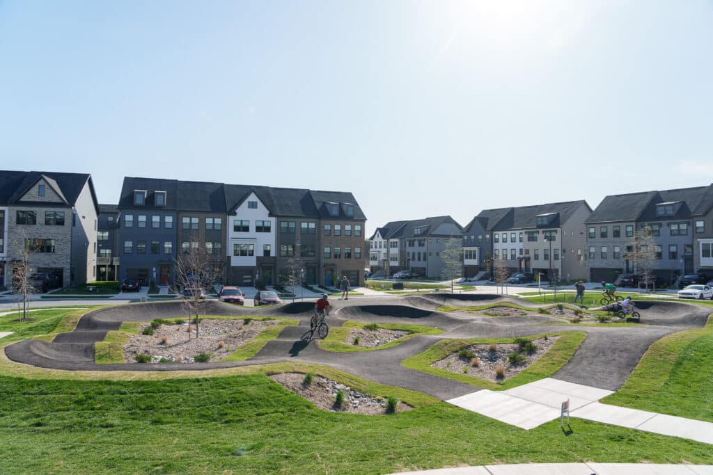 A pump track with several cyclists is set in a residential neighborhood. Modern, multi-story homes with dark roofs line the background. The sun shines brightly, casting soft shadows on the green grass and winding paths.