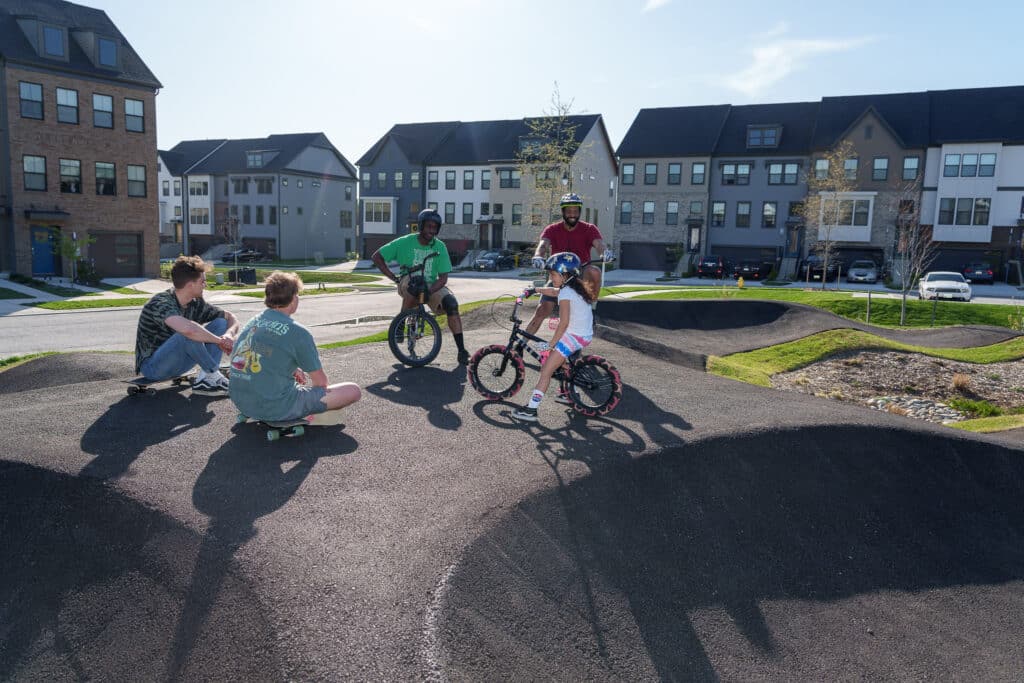 A group of people gather on bikes and a skateboard at a paved pump track in a residential area. The sunny day highlights colorful houses and lush greenery in the background. A child on a bike wears a helmet, engaging in conversation with the group.