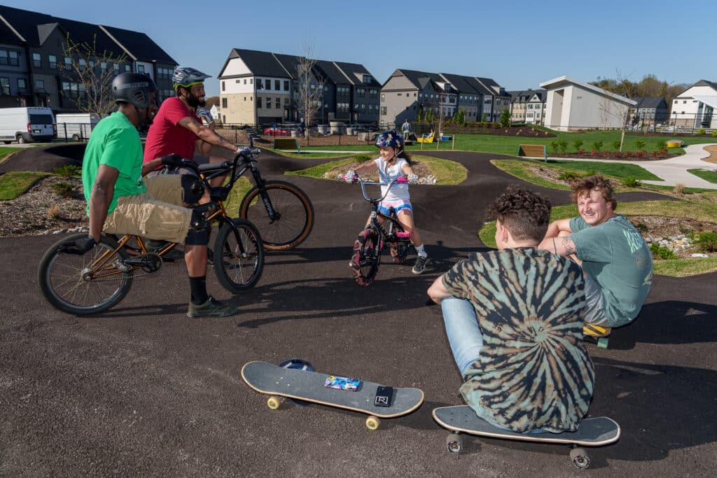 A group of people enjoy a sunny day at a skate park. Two men stand with bicycles, a young girl rides a bike, and two people sit on the pavement with skateboards. Residential buildings and green spaces are in the background.