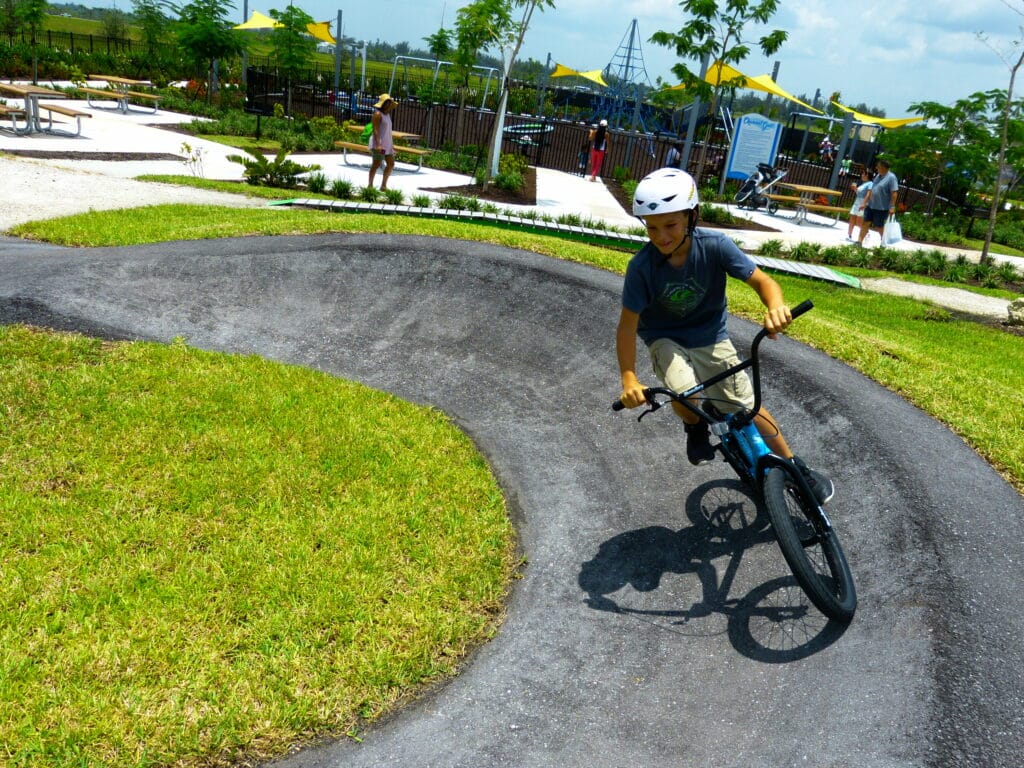 A child wearing a helmet rides a BMX bike along a curved track in a park. The park features lush grass, trees, and a playground in the background with adults and children enjoying the space.
