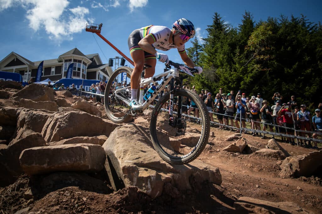 A cyclist in professional gear navigates a rocky mountain trail during a race. Spectators watch from the sidelines against a backdrop of trees and a building. The cyclist displays intense focus and control.