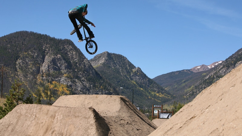 A person on a BMX bike performs a mid-air trick over a dirt jump against a backdrop of mountainous terrain. The sky is clear and blue, highlighting the natural scenery and the bikers skill.