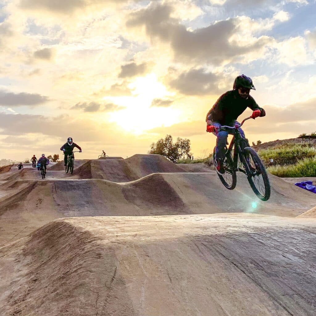 Cyclists ride on a dirt bike trail at sunset, with the golden sky casting long shadows. The foreground features a rider actively jumping over a hill, followed by others in the distance, showcasing the dynamic outdoor scene.