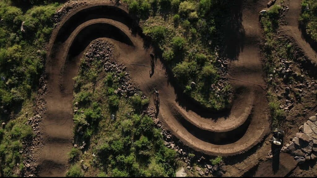Aerial view of a winding dirt trail with sharp curves through a grassy landscape. Rocks and patches of greenery surround the well-trodden path, providing a natural setting.
