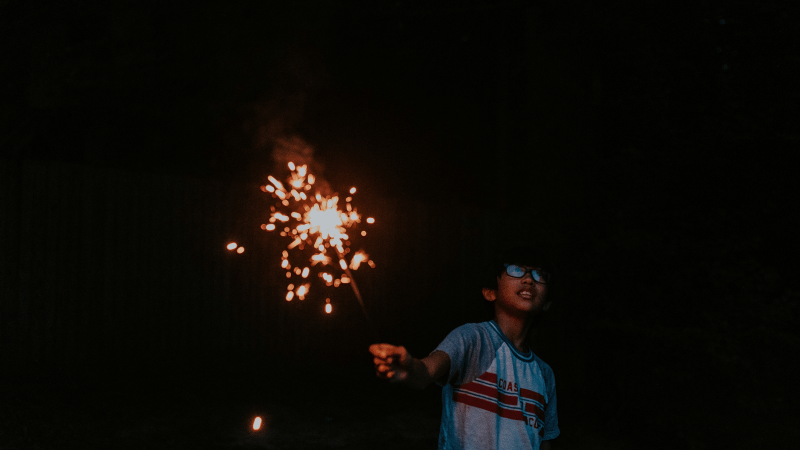 A young boy wearing glasses holds a lit sparkler at night. The sparkler creates bright, glowing sparks that illuminate his face and a small area around him, contrasting against the dark background.