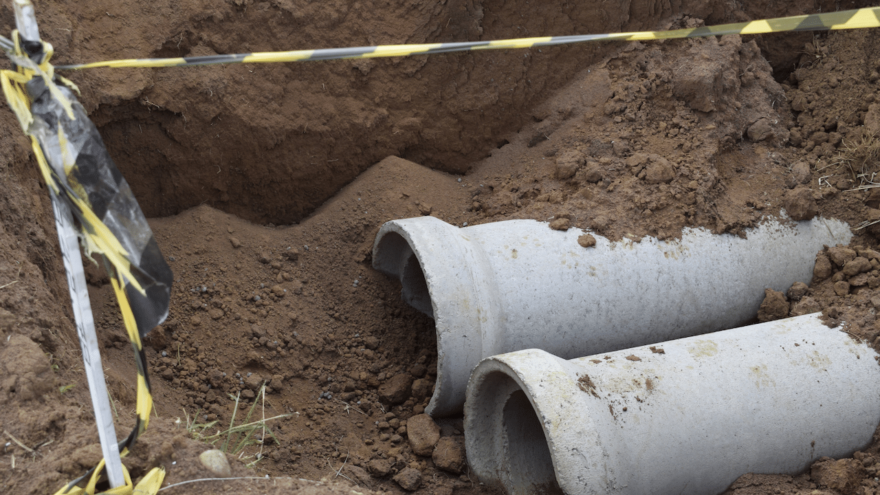Two concrete pipes partially buried in a dirt excavation site, surrounded by loose soil. Yellow and black caution tape is visible, marking the area.