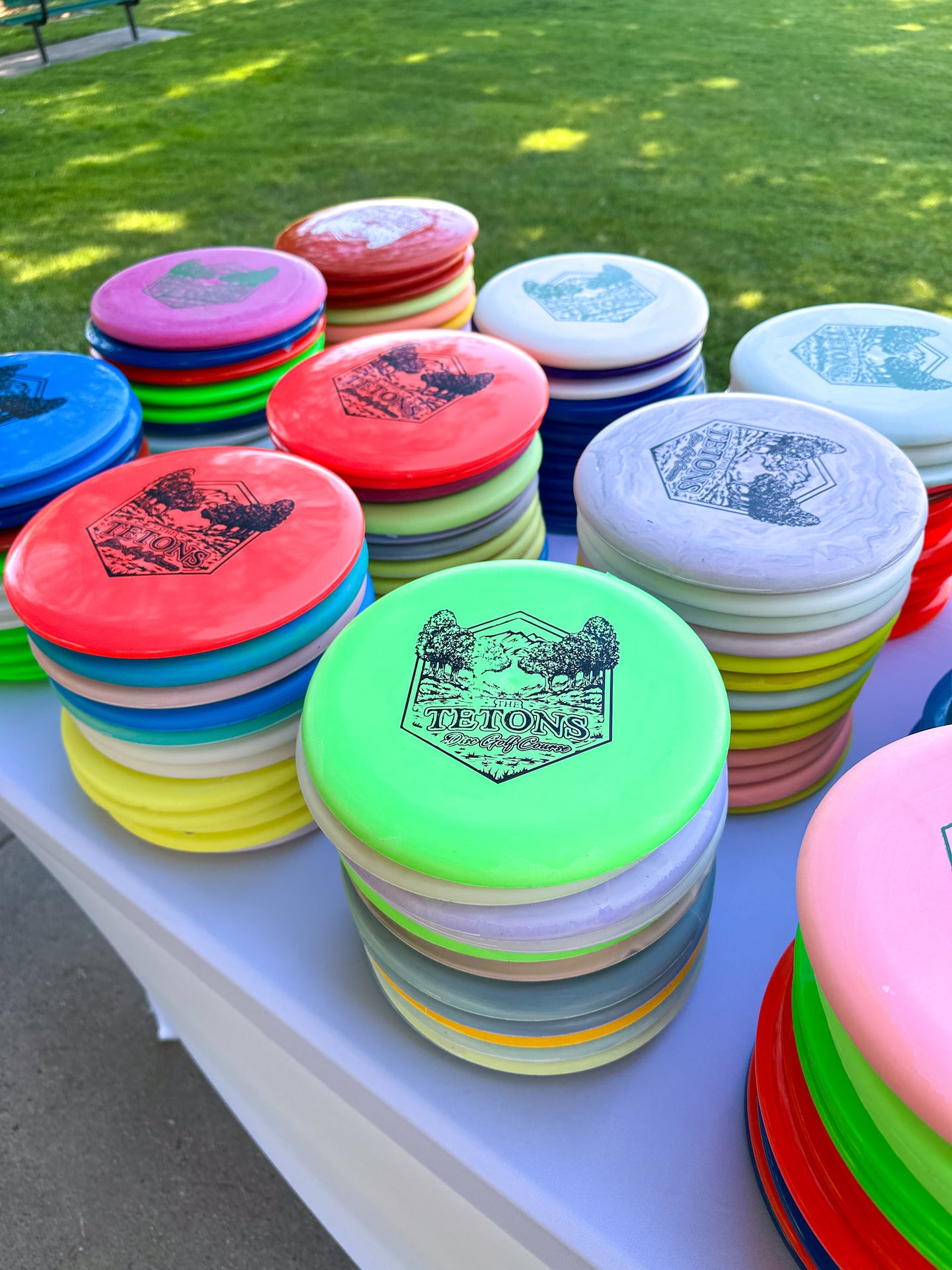 Stacks of colorful frisbee golf discs with various designs displayed on a white table outdoors. A grassy area is visible in the background.