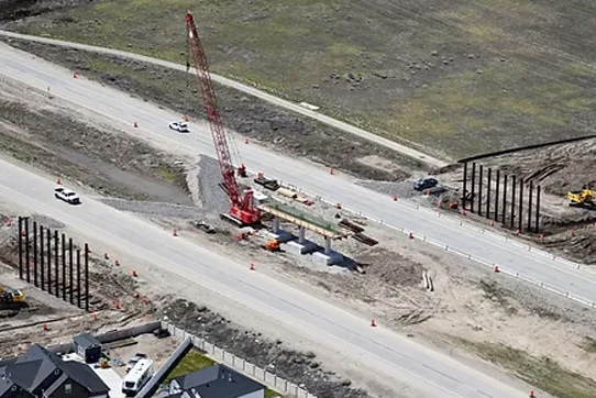 Aerial view of a construction site with a red crane on a road, surrounded by construction materials and vehicles. Metal beams are being installed, with traffic cones outlining the area. A grassy field and residential houses are visible nearby.