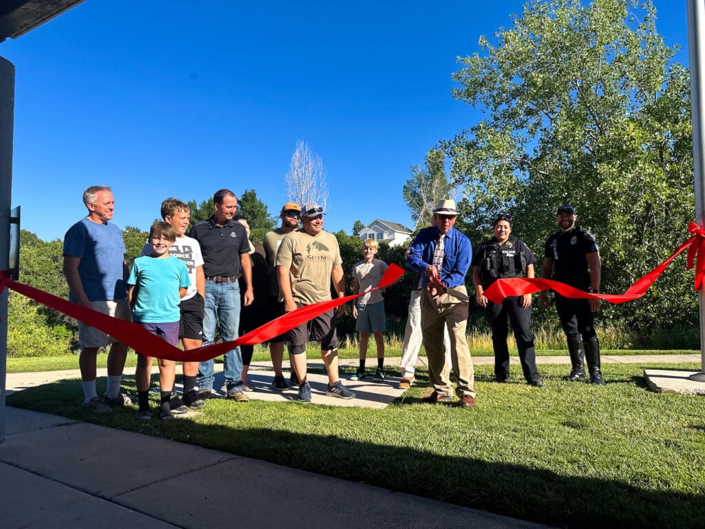 A group of people, including adults and children, stand outdoors on grass for a ribbon-cutting ceremony. A man holds large scissors to cut a red ribbon while others watch and smile. Trees and a clear blue sky are in the background.